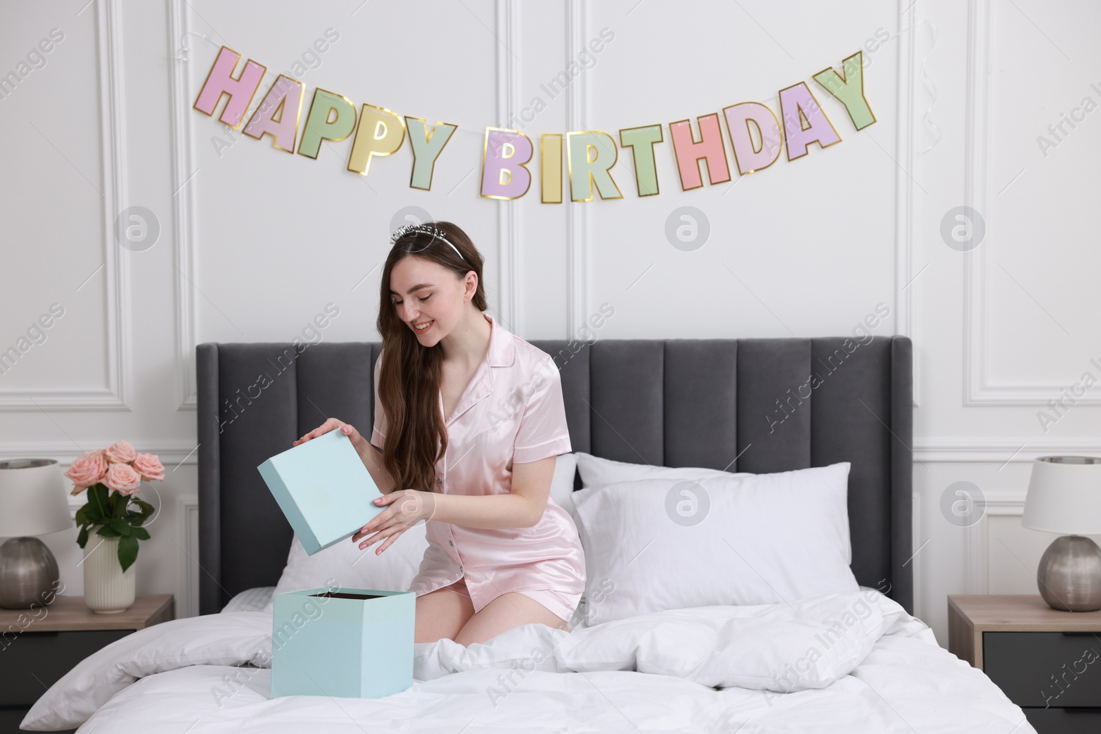 Photo of Beautiful young woman with headband opening gift box on bed in room. Happy Birthday