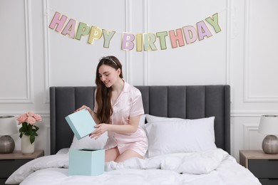 Beautiful young woman with headband opening gift box on bed in room. Happy Birthday