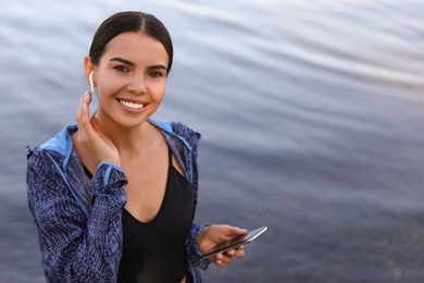 Photo of Young sportswoman with wireless earphones and smartphone near river