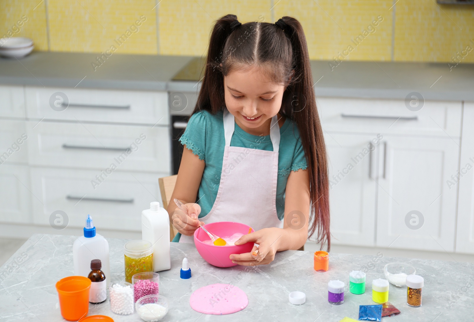 Photo of Cute little girl mixing ingredients with silicone spatula at table in kitchen. DIY slime toy