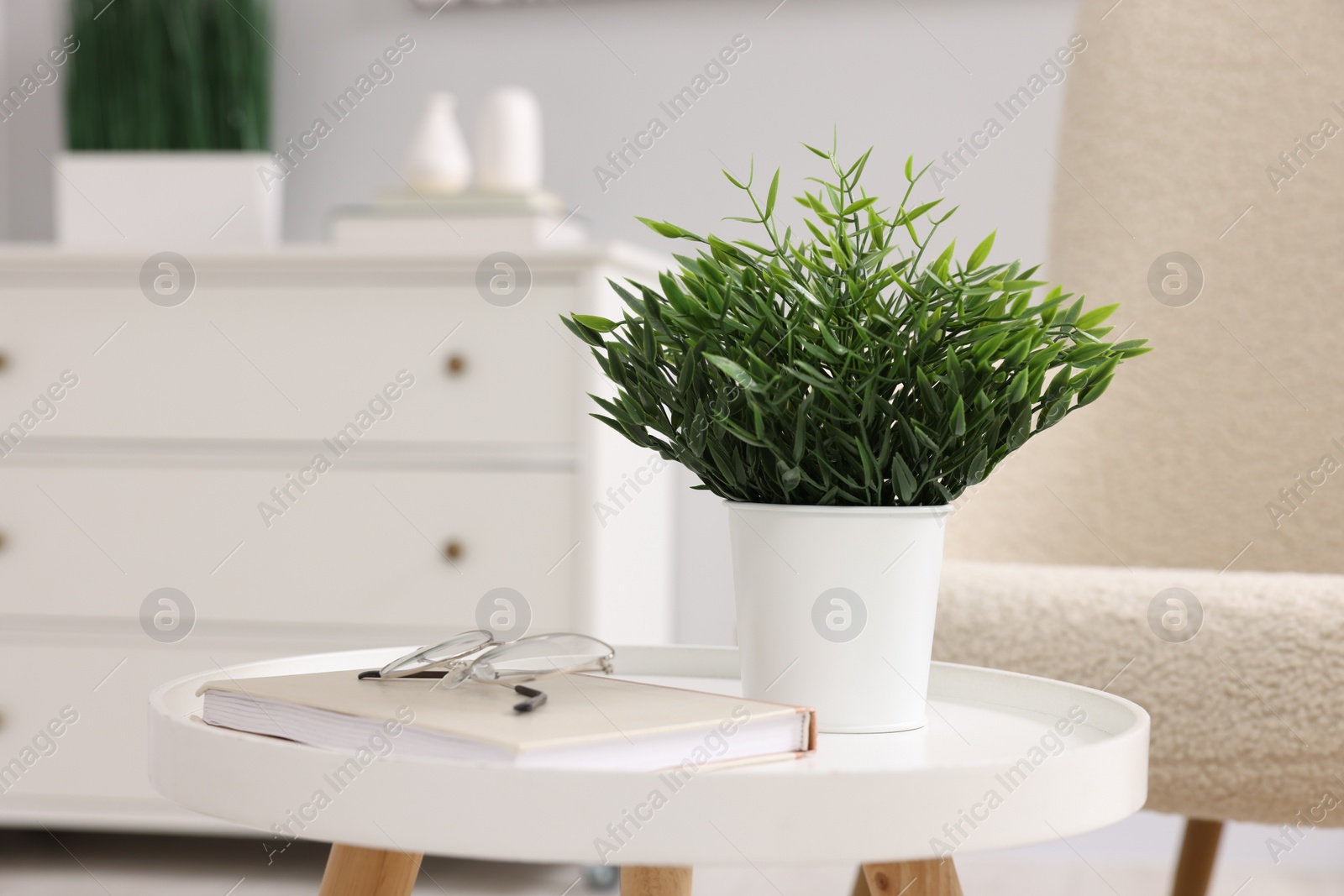 Photo of Potted artificial plant, book and glasses on side table near armchair indoors