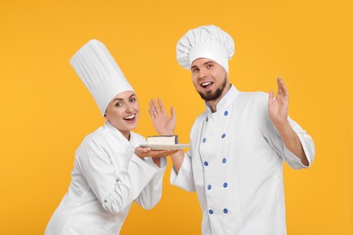 Photo of Happy professional confectioners in uniforms holding delicious cheesecake on yellow background