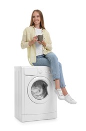 Beautiful young woman with cup of drink on washing machine with laundry against white background