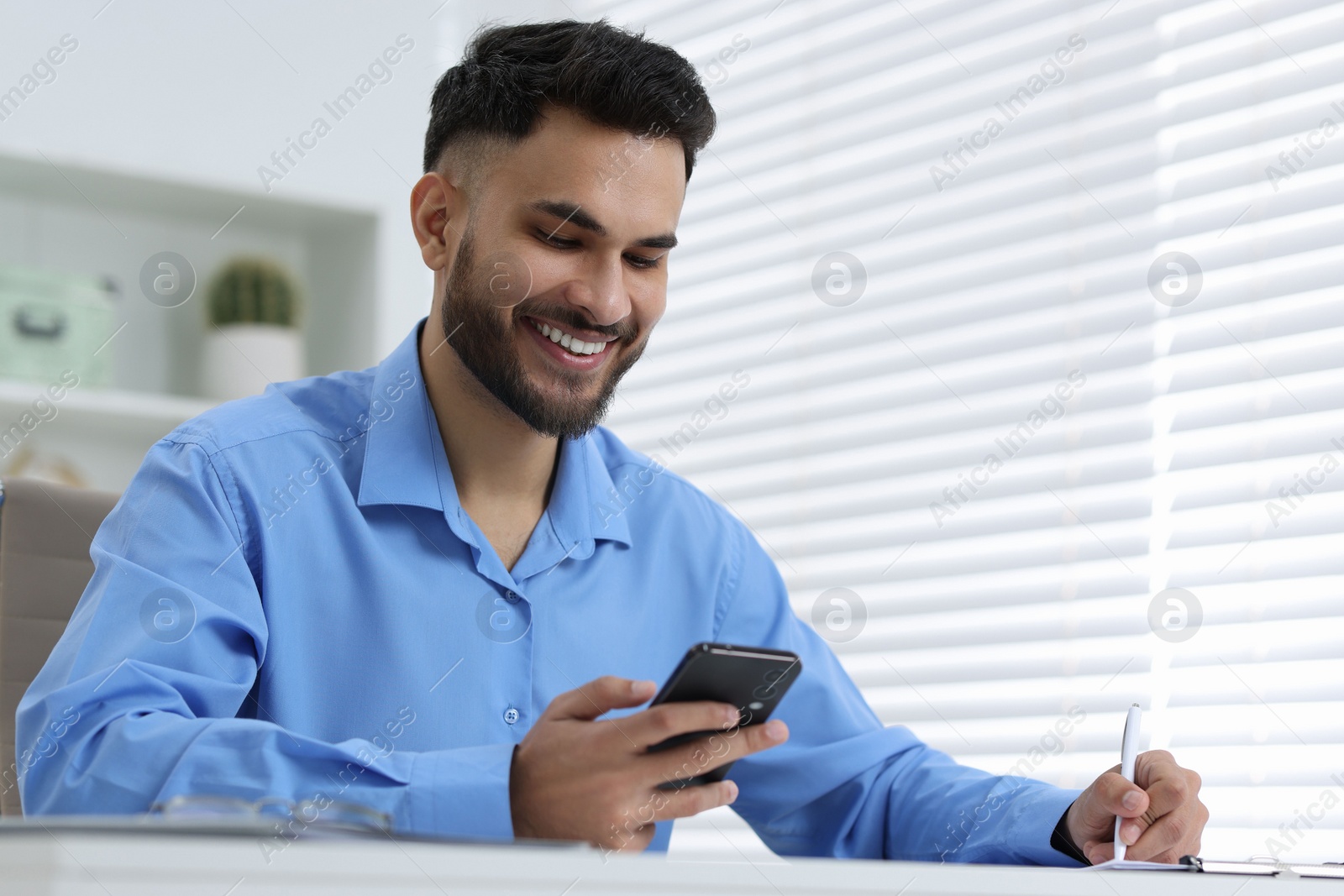 Photo of Happy young man using smartphone while working at white table in office, low angle view