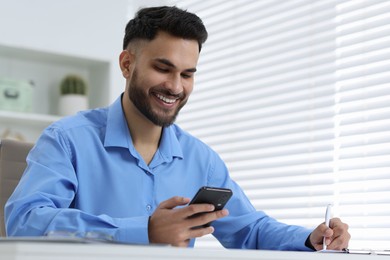 Photo of Happy young man using smartphone while working at white table in office, low angle view