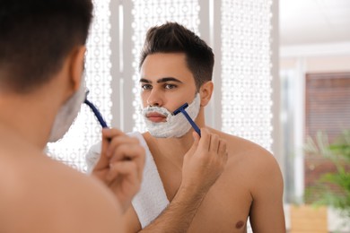 Handsome young man shaving with razor near mirror in bathroom