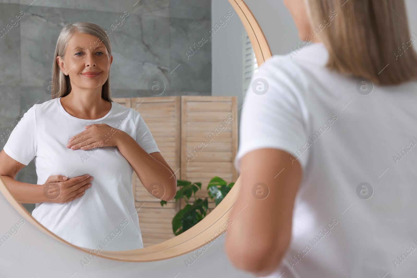 Photo of Beautiful senior woman doing breast self-examination near mirror in bathroom