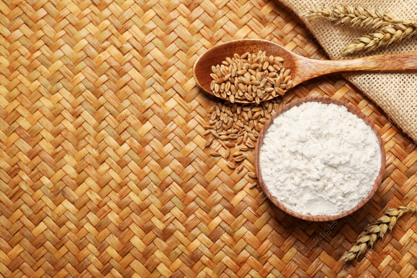 Photo of Wheat flour in bowl, spikes and grains on wicker mat, flat lay. Space for text