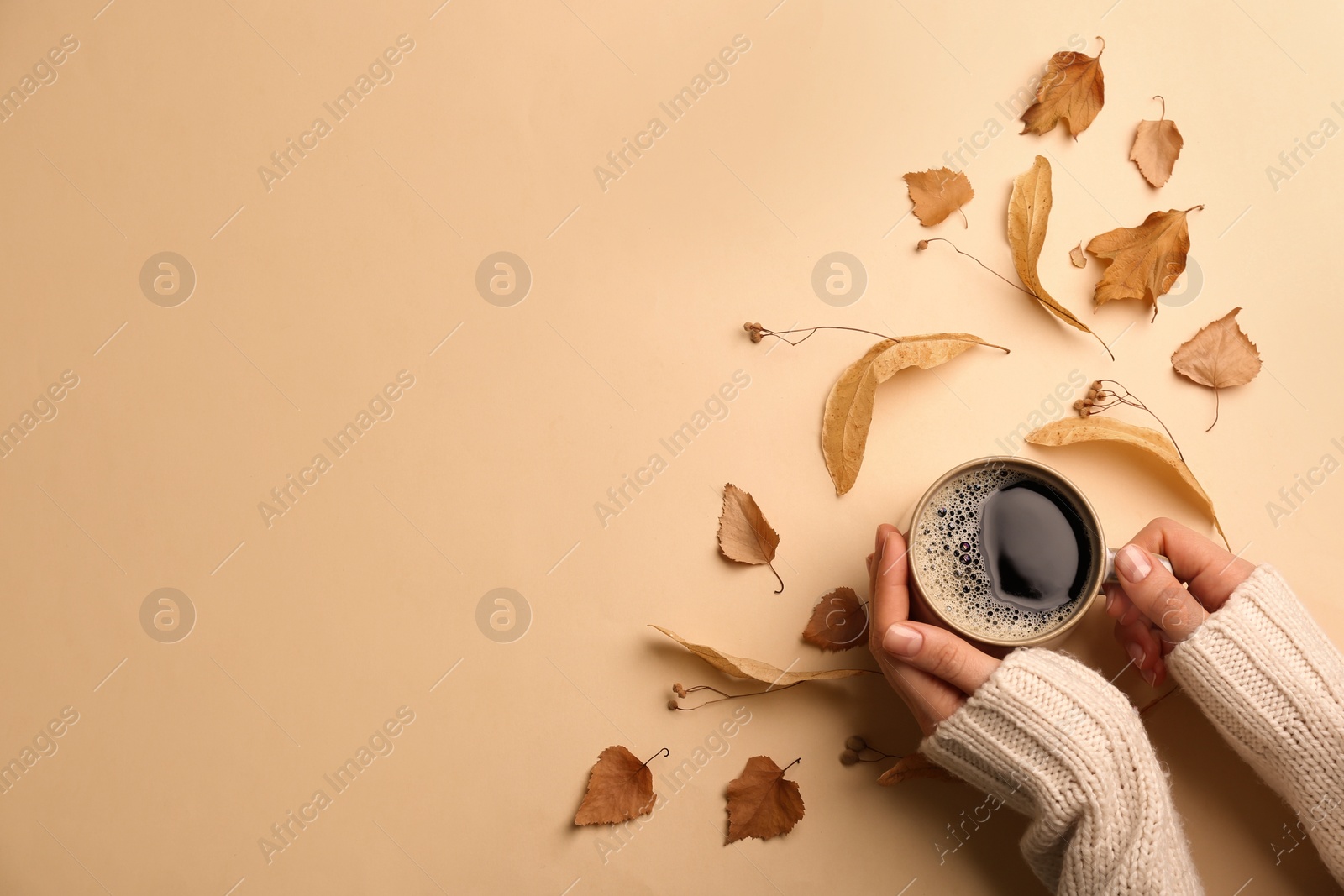 Photo of Woman with cup of hot drink surrounded by autumn leaves on beige background, space for text. Cozy atmosphere