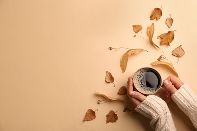 Photo of Woman with cup of hot drink surrounded by autumn leaves on beige background, space for text. Cozy atmosphere