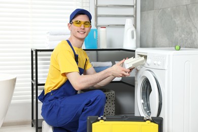 Smiling plumber wearing protective glasses repairing washing machine in bathroom