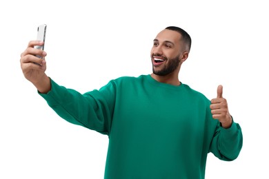 Smiling young man taking selfie with smartphone and showing thumbs up on white background