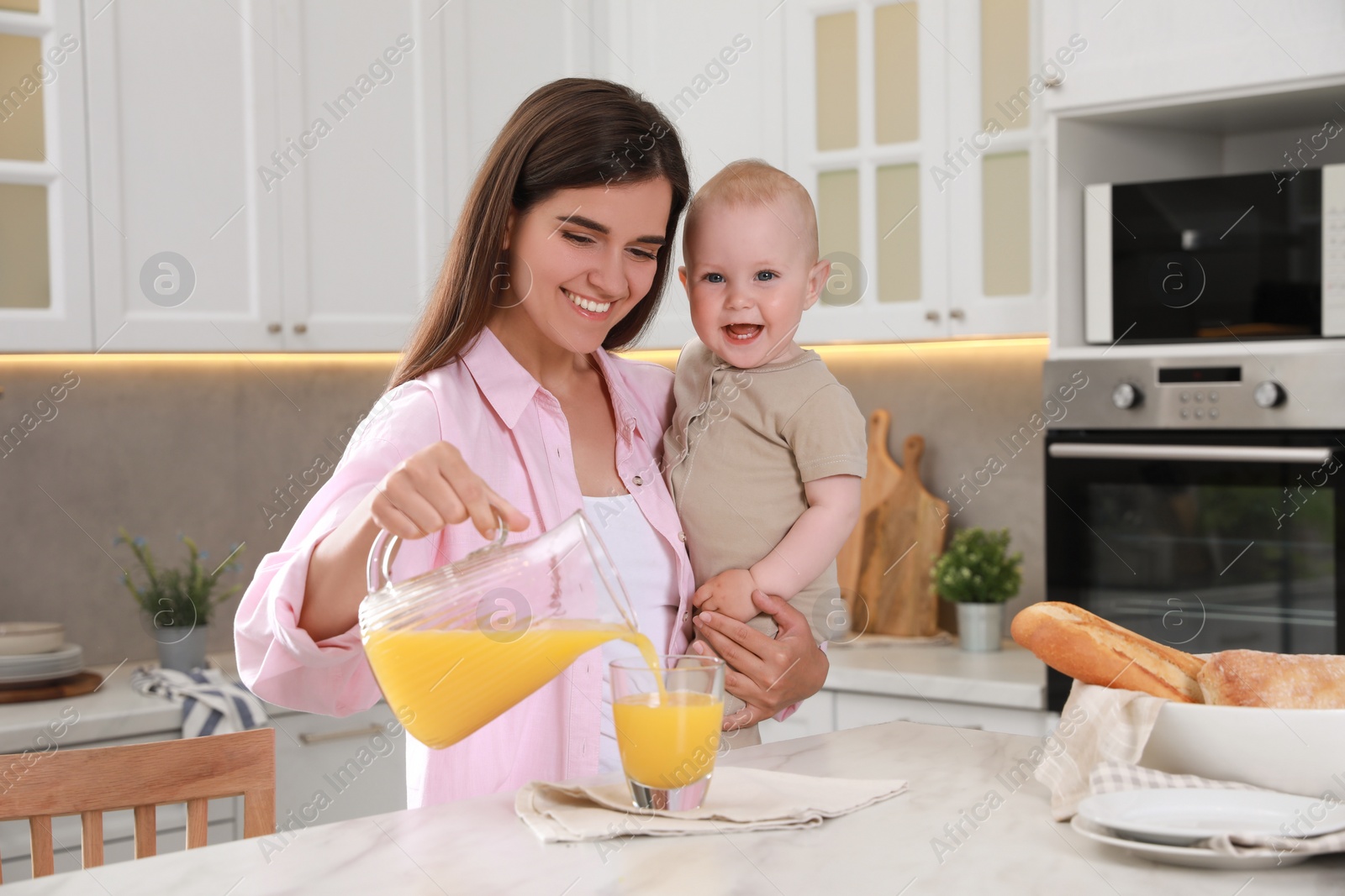 Photo of Happy young woman holding her cute little baby while pouring juice into glass in kitchen