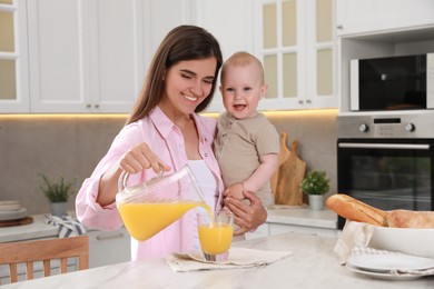Happy young woman holding her cute little baby while pouring juice into glass in kitchen