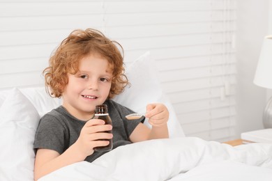 Photo of Cute boy holding measuring spoon with cough syrup in bed. Effective medicine