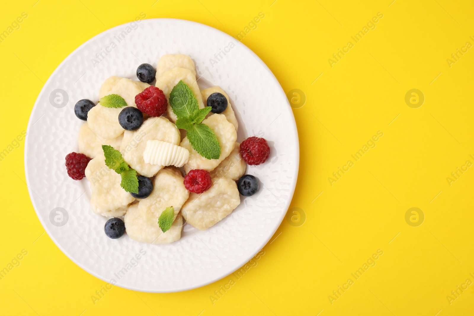 Photo of Plate of tasty lazy dumplings with berries, butter and mint leaves on yellow background, top view. Space for text