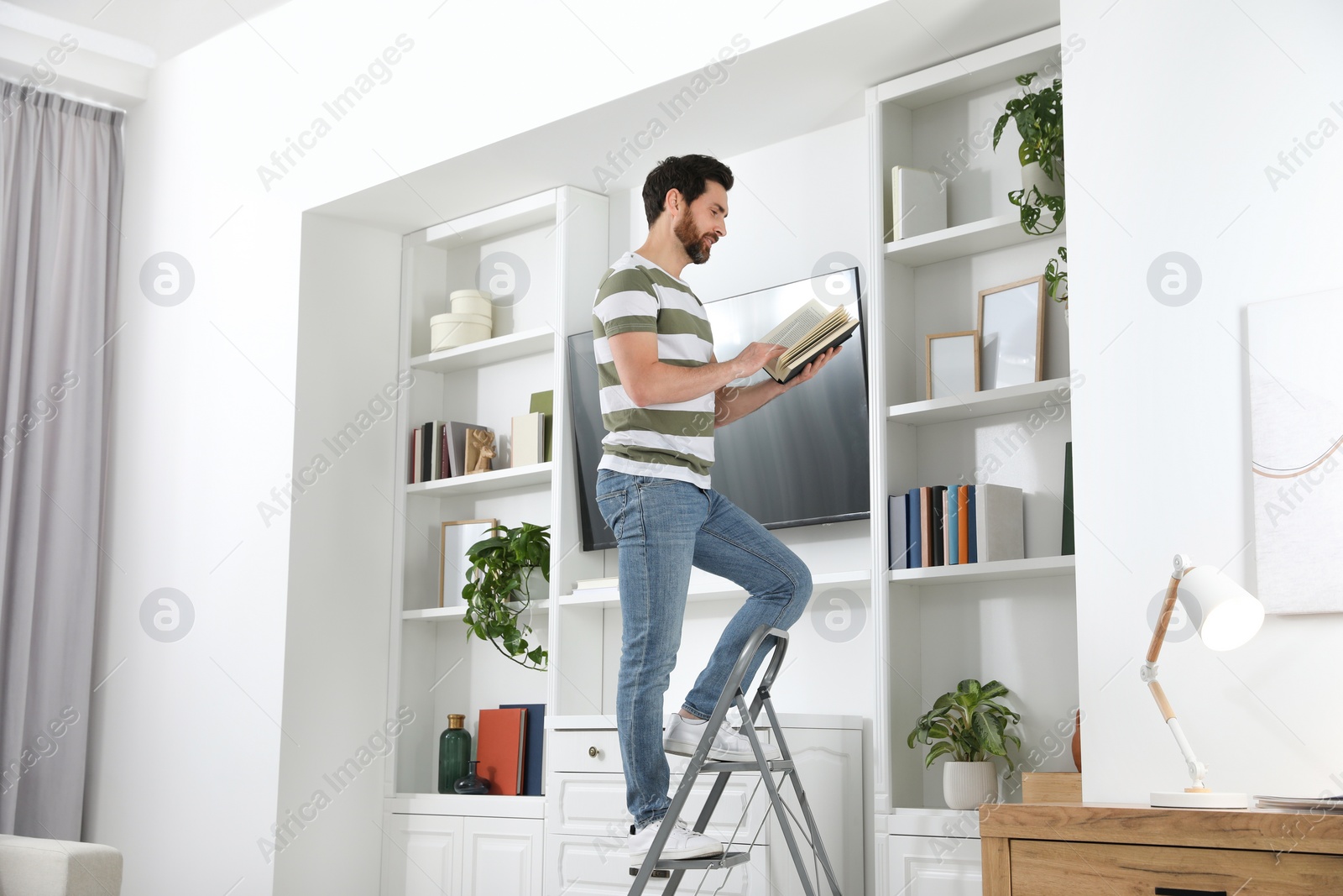 Photo of Man reading book on metal folding ladder at home