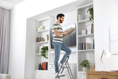 Photo of Man reading book on metal folding ladder at home