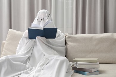 Photo of Creepy ghost. Person covered with white sheet reading book on sofa at home