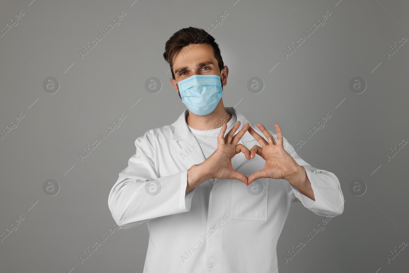 Photo of Doctor in protective mask making heart with hands on grey background