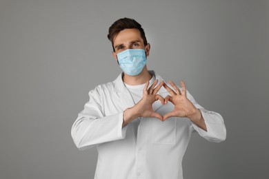 Photo of Doctor in protective mask making heart with hands on grey background