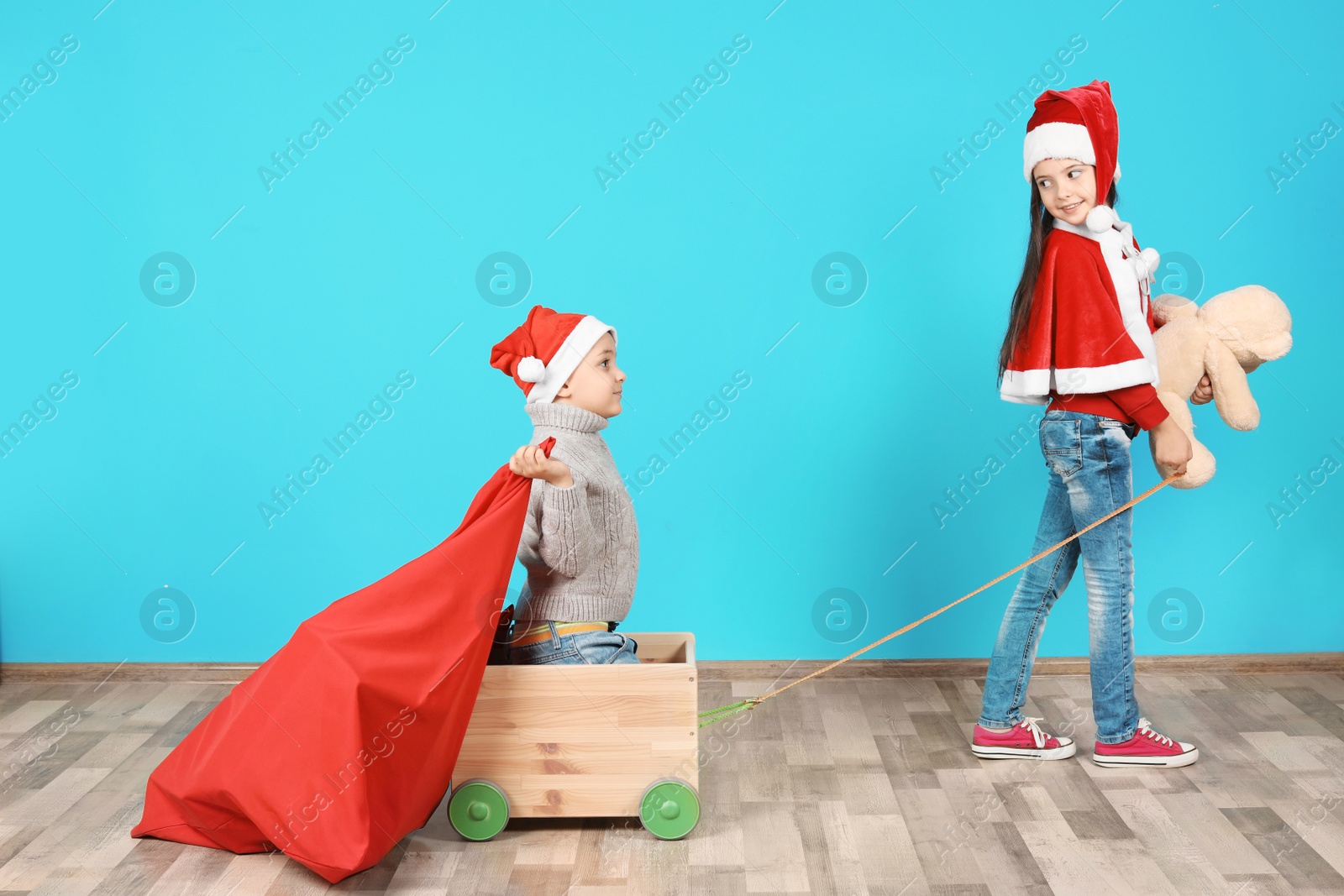 Photo of Cute little children in Santa hats playing with toy cart and Christmas sack near color wall