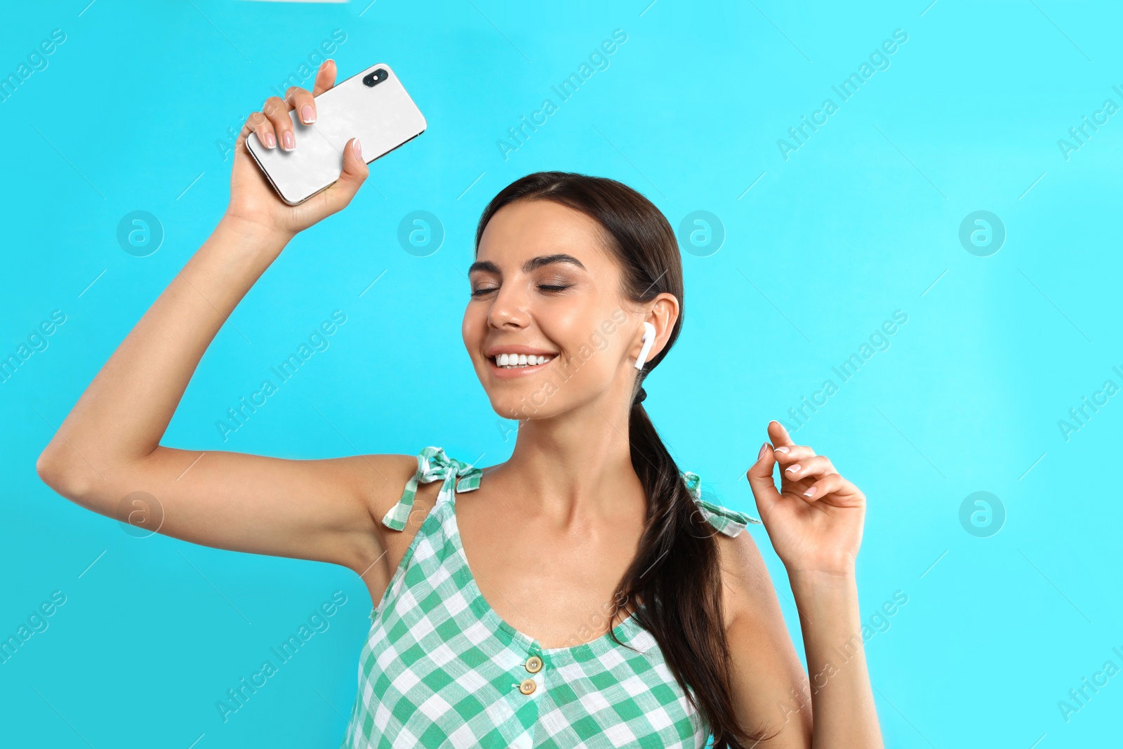 Photo of Happy young woman with smartphone listening to music through wireless earphones on light blue background