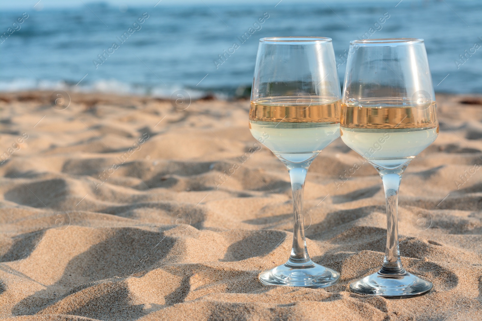 Photo of Glasses of tasty wine on sand near sea, space for text