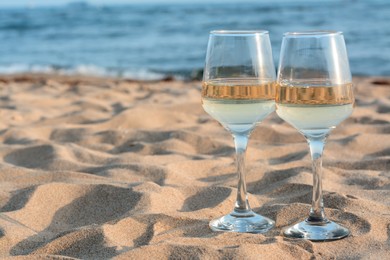Glasses of tasty wine on sand near sea, space for text