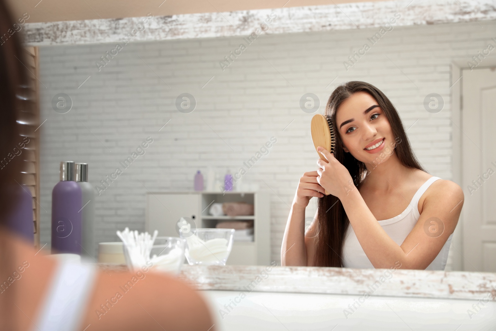 Photo of Beautiful young woman with hair brush looking into mirror in bathroom