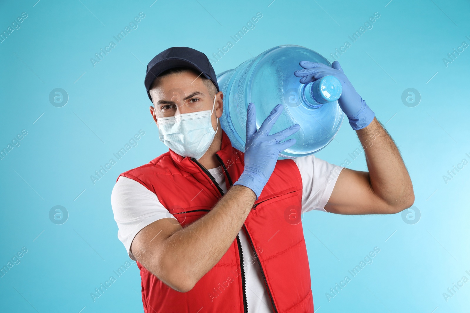 Photo of Courier in face mask with bottle of cooler water on light blue background. Delivery during coronavirus quarantine