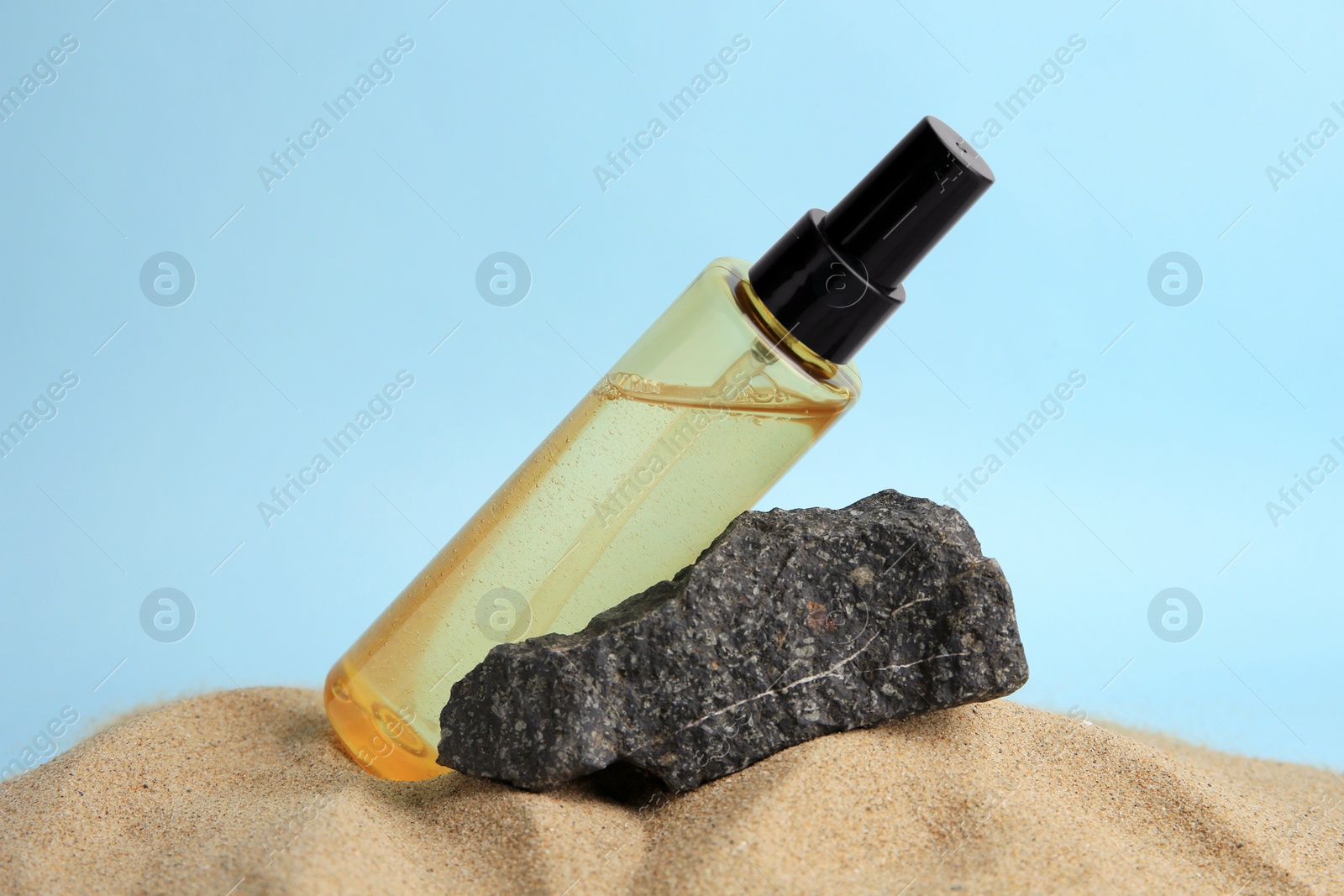 Photo of Bottle with serum and stone on sand against light blue background, closeup. Cosmetic product