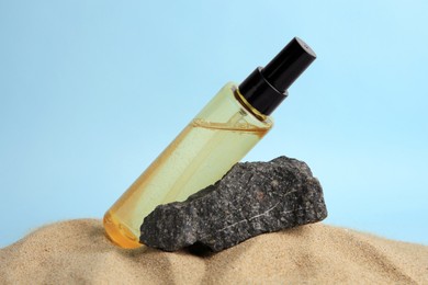 Bottle with serum and stone on sand against light blue background, closeup. Cosmetic product