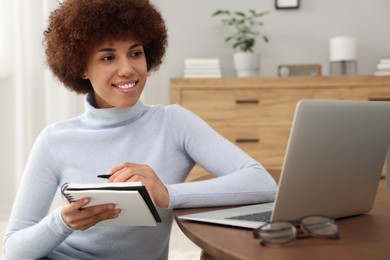 Photo of Beautiful young woman using laptop and writing in notebook at wooden coffee table in room