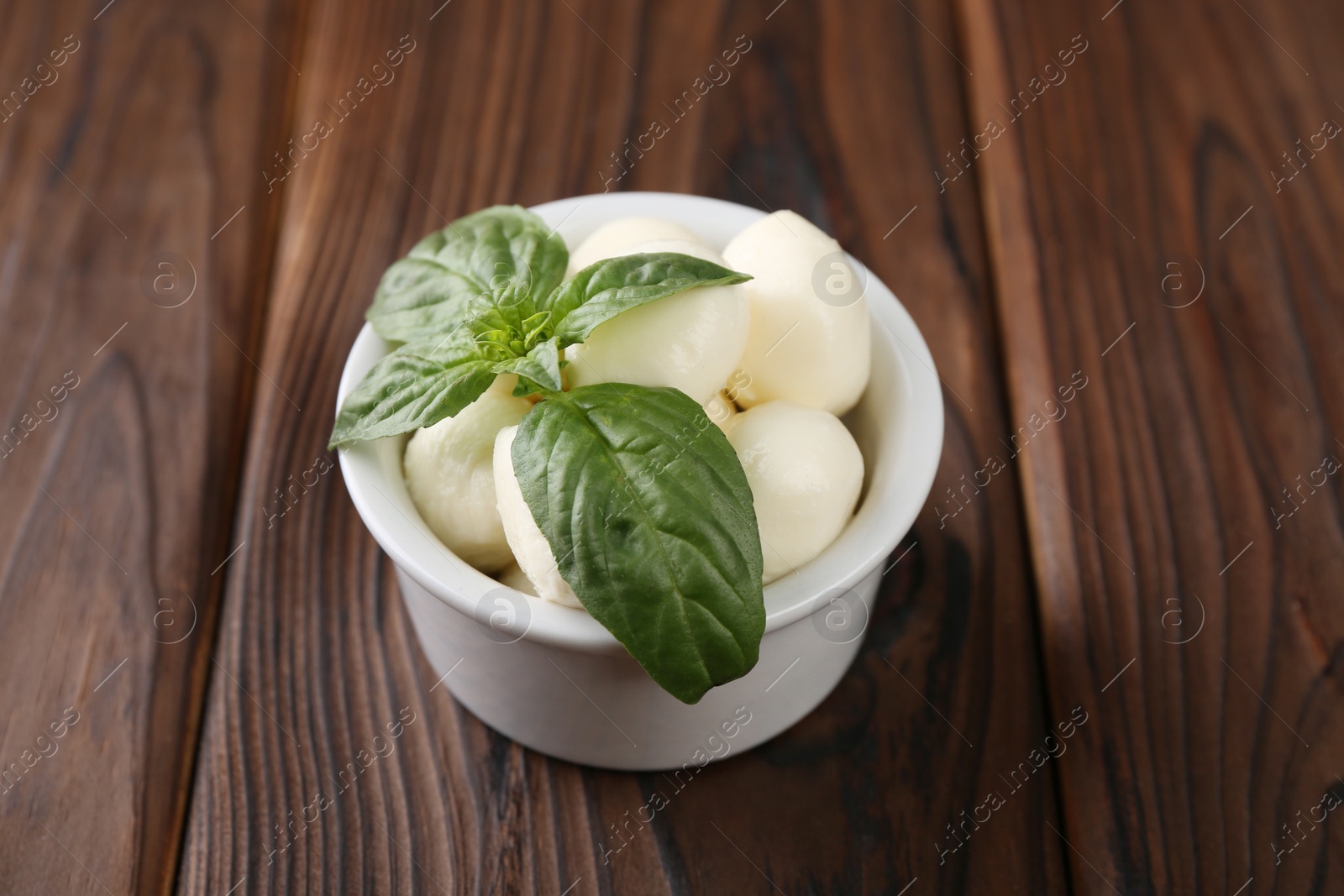 Photo of Tasty mozarella balls and basil leaves in bowl on wooden table, closeup