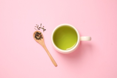 Photo of Spoon with green tea leaves and cup of hot beverage on pink background, flat lay