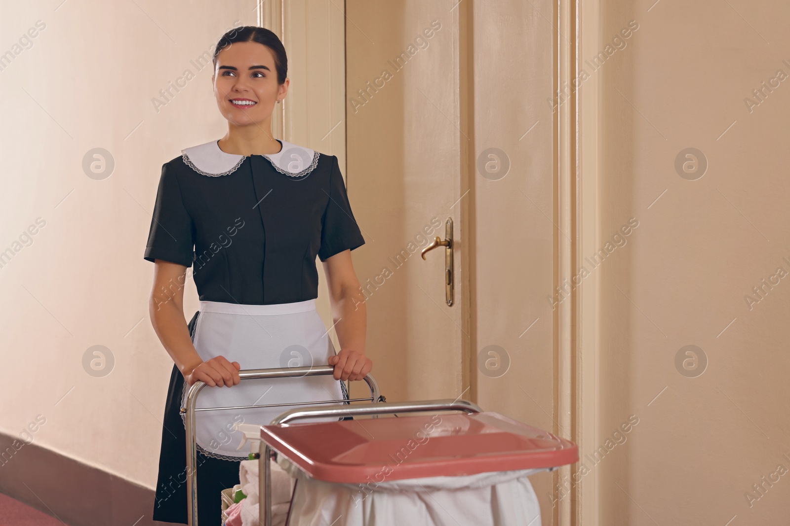 Photo of Young chambermaid with cart and cleaning products in hotel. Space for text