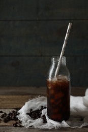 Delicious iced coffee in glass bottle with straw near beans on wooden table. Space for text