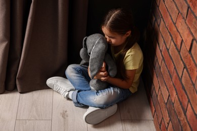 Child abuse. Upset little girl with toy bunny sitting on floor near brick wall indoors