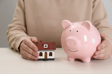 Photo of Man with house model and piggy bank at wooden table, closeup. Saving money concept