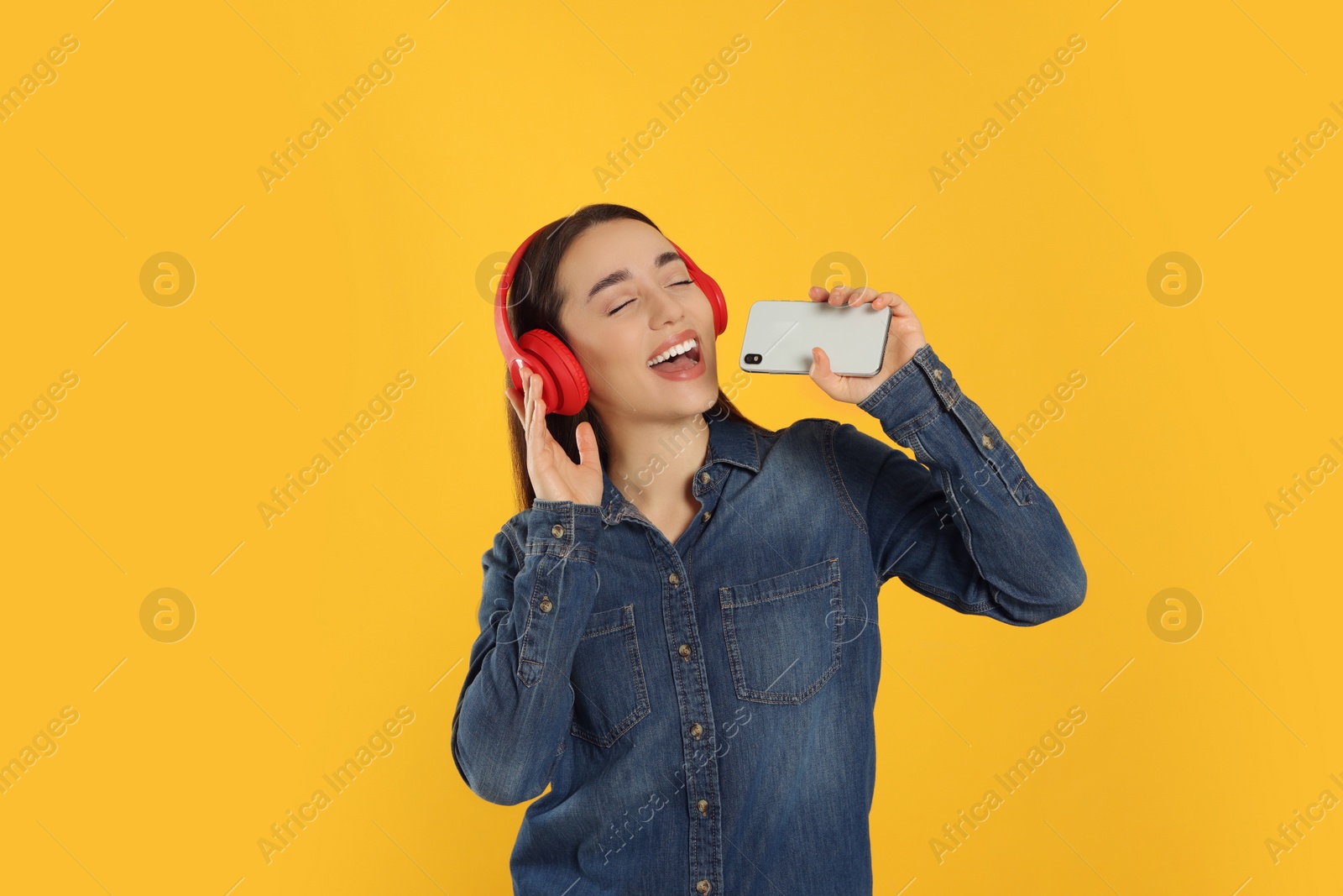 Photo of Happy woman in headphones enjoying music and singing into smartphone on orange background