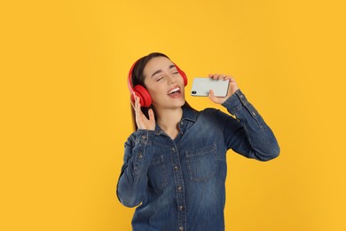 Photo of Happy woman in headphones enjoying music and singing into smartphone on orange background