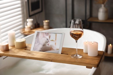 Photo of Wooden tray with tablet, wine and candles on bathtub in bathroom