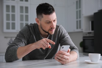Photo of Emotional man with smartphone at table in kitchen. Online hate concept