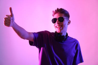 Photo of Young man with sunglasses showing thumbs up on pink background in neon lights