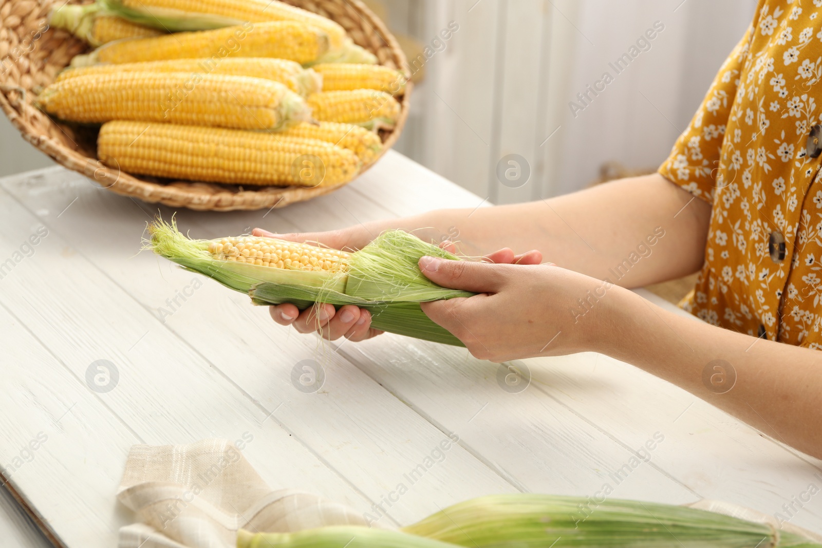 Photo of Woman husking corn cob at white wooden table, closeup