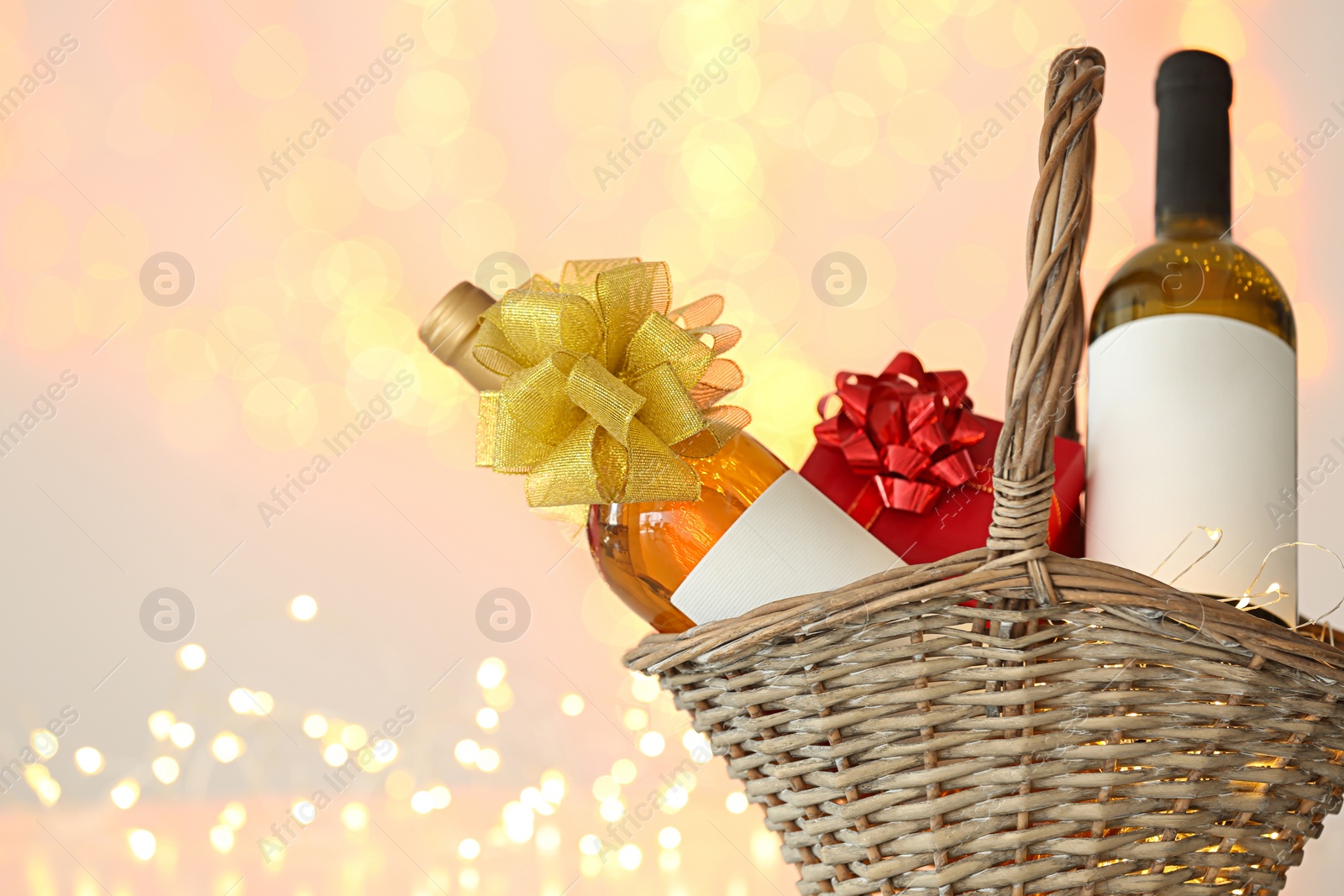 Photo of Wicker basket with bottles of wine and gift box against blurred lights. Space for text