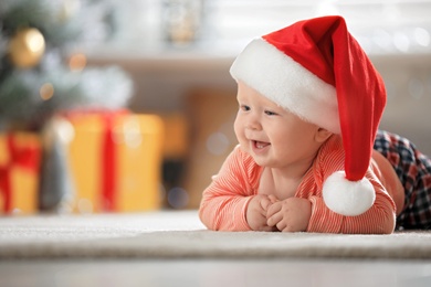 Photo of Little baby wearing Santa hat on floor indoors. First Christmas