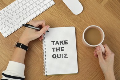 Woman with cup of coffee writing phrase Take the quiz in notebook at wooden table, top view