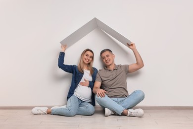 Young family housing concept. Pregnant woman with her husband sitting under cardboard roof on floor indoors